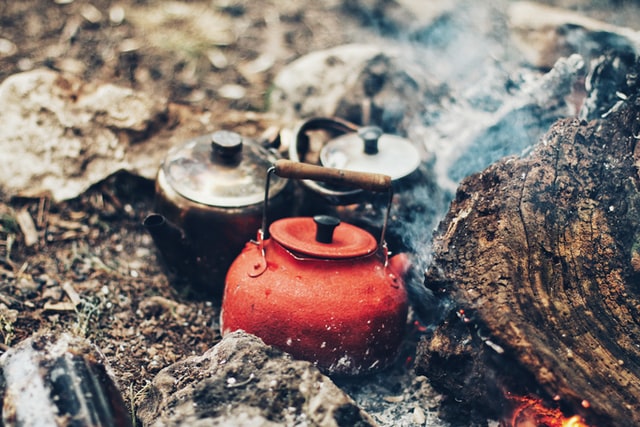 three boiling water filled kettles of multiple colors placed near a campfire to keep warm